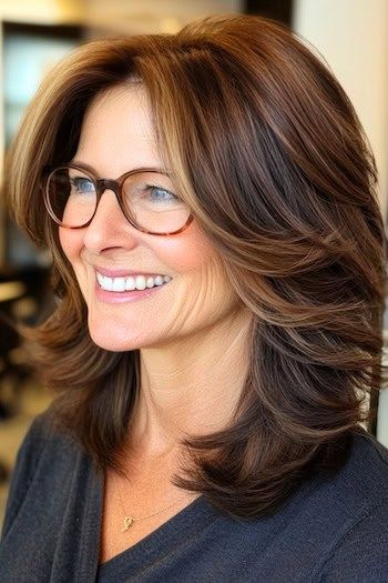 Peinado con capas de plumas en una mujer sonriente de unos 50 años con pelo castaño y gafas, vista lateral.